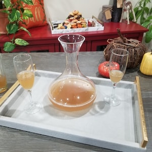 Peut inclure: Un plateau de service gris avec des accents dorés contient une carafe en verre transparent remplie d'un liquide brun clair. Deux verres à pied transparents remplis du même liquide sont sur le plateau.