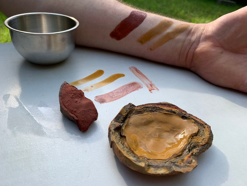 May include: A close-up of a variety of natural pigments, including red, brown, and yellow, displayed on a white surface. The pigments are shown in both their raw form and as swatches on a person's arm.