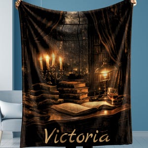 May include: A dark brown blanket featuring a cozy library scene with a lit candelabra, books, a lamp, and a glass of wine. The name "Victoria" is printed in gold script at the bottom. The blanket is held up by a person.