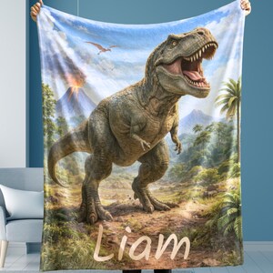 May include: A plush blanket featuring a detailed illustration of a Tyrannosaurus Rex in a prehistoric landscape with a volcano and palm trees. The blanket is light blue with the name "Liam" in a soft, beige font.