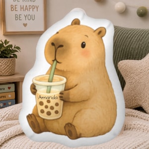 May include: A plush, light brown capybara-shaped pillow holding a bubble tea. The bubble tea cup is clear with brown boba and a green straw. The name "Amanda" is printed on the cup. The pillow is on a white background.