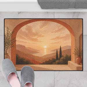 May include: Rectangular floor mat featuring a scenic landscape with an archway, mountains, and a sunset over the water. The mat has a black border and is in warm tones of orange, brown, and beige. Two gray slippers with pink interiors are in the foreground.