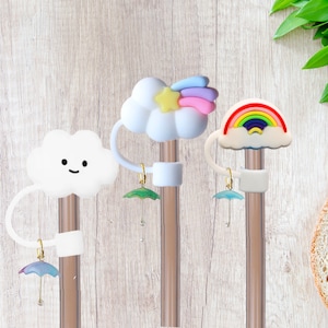 Puede incluir: Tres tapones de paja coloridos con un diseño de nube y arcoíris. Cada tapón tiene un pequeño amuleto de paraguas colgante.