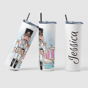 May include: Three white stainless steel tumblers with a Christmas theme. The center tumbler features a watercolor illustration of a Nutcracker holding a gift box. The tumbler on the left has the same illustration but is cropped to show only the Nutcracker. The tumbler on the right has the name "Jessica" written in black script.