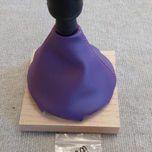 May include: Purple leather gear shift boot with a black gear shift knob with a silver top. The knob has the numbers 1, 2, 3, 4, 5, and R on it. The boot is sitting on a wooden base. A small plastic bag with a spring and a screw is in the lower right corner of the image.