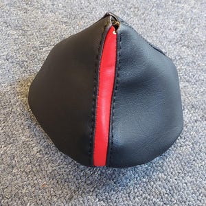 Könnte beinhalten: Schwarzes Lederobjekt mit einem vertikalen roten Streifen. Das Objekt hat eine abgerundete Form und scheint eine Abdeckung oder ein Zubehör zu sein. Der Artikel befindet sich auf einer grauen Oberfläche.