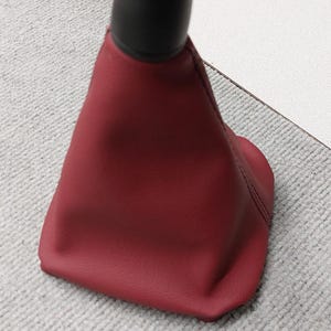 Puede incluir: Una funda de palanca de cambios de cuero rojo con una parte superior de plástico negro. La funda está diseñada para ajustarse a una palanca de cambios.