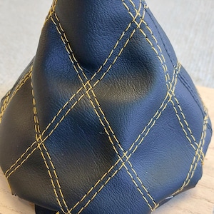 Peut inclure: Pommeau de levier de vitesse en cuir noir avec surpiqûres jaunes en forme de losange. Le pommeau est sur une base en bois.