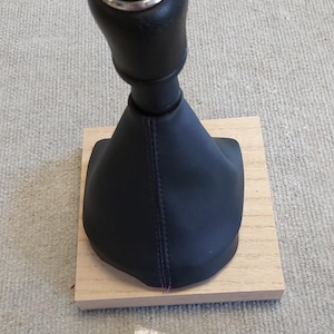 May include: Black leather gear shift knob with a silver trim and a 5-speed gear pattern. The knob is attached to a black leather boot and is sitting on a wooden base. A small bag containing a spring and a screw is in the foreground.