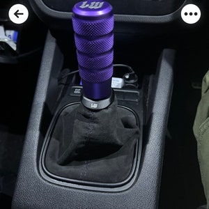 Puede incluir: Pomo de cambio de marchas de coche morado y negro con una funda de cuero negro. El pomo tiene un logotipo "LW" en relieve en la parte superior.