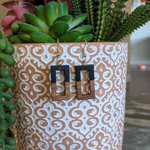 May include: A pair of wooden earrings with black and brown rectangular shapes. The earrings are hanging from gold hooks.