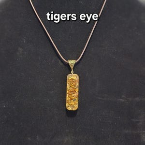 May include: A gold-colored pendant necklace with a rectangular tiger's eye stone. The pendant is suspended from a brown cord. The text "tigers eye" is displayed above the necklace.