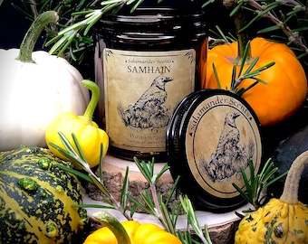 Könnte beinhalten: Ein braunes Glasgefäß mit einer Kerze und einem schwarz-weißen Etikett mit der Aufschrift "Salamander Scents Samhain Pumpkin Spice" mit einem Raben und Kürbissen. Das Kerzengefäß ist von Kürbissen und Rosmarin umgeben.