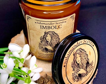 Könnte beinhalten: Ein braunes Glas-Kerzengefäß mit einer weißen Kerze darin. Das Gefäß hat ein Etikett mit der Aufschrift "Salamander Scents" und "IMBOLC" in schwarzer Schrift. Das Etikett zeigt auch eine Zeichnung einer Frau, die einen Zweig Grünzeug hält. Die Kerze ist mit "Lemongrass" und "hand poured soy" beschriftet. Das Gefäß steht auf einer Holzoberfläche mit einem Strauß weißer Schneeglöckchen.
