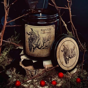 Op de afbeelding: Een bruine glazen kaars pot met een zwarte raaf illustratie en de tekst "RAVEN Oud & Vanilla" op het etiket. De kaars is handgegoten en gemaakt van sojawas.