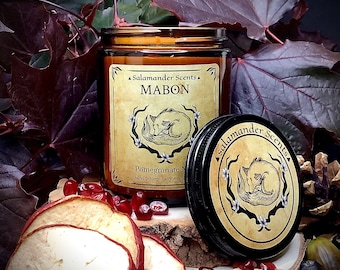 Könnte beinhalten: Ein braunes Glasgefäß mit einer Kerze und einem goldenen Etikett mit der Aufschrift "Salamander Scents Mabon Pomegranate Moon hand poured soy wax". Das Etikett zeigt auch eine stilisierte Illustration eines schlafenden Fuchses. Das Kerzengefäß steht auf einer Holzoberfläche mit getrockneten Apfelscheiben und Granatapfelkernen.