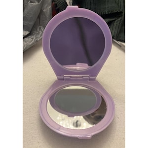 May include: A lavender compact mirror, open to reveal two reflective surfaces. The compact has a rounded, oval shape and a hinged design. The mirrors are set within a matching lavender frame.