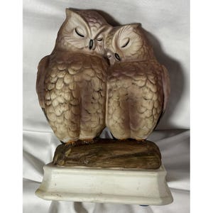 May include: A ceramic figurine of two owls, nestled together, with closed eyes. The owls are light brown with textured feathers, perched on a faux wooden branch atop a white base. A decorative item.