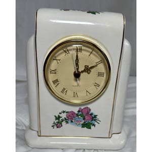 May include: A white ceramic quartz clock with a gold-tone clock face and Roman numerals. The clock features a floral design with pink and blue roses. Gold trim accents the edges of the clock.