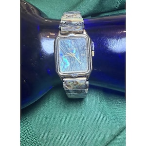 May include: A silver-toned wristwatch with a rectangular face and a matching band. The watch face has a shimmering, iridescent blue-green surface. The watch hands are gold-toned, and the watch is set against a dark blue background.