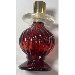 May include: A decorative, ruby-red glass perfume bottle with a ribbed, bulbous body and a clear, circular atomizer. The bottle has a gold-colored cap and a flared base. The design is vintage and elegant.