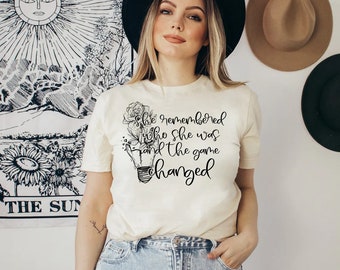 May include: A white t-shirt with a black graphic design featuring a lightbulb with a flower growing out of it. The text on the shirt reads "She remembered who she was and the game changed."