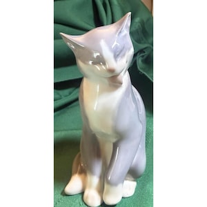 May include: A porcelain cat figurine in a seated position, featuring a smooth, glossy finish with soft lavender-grey and white color blocking. The cat is depicted with its head tilted and tongue slightly visible, showcasing a clean, minimalist aesthetic.