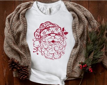 May include: White t-shirt featuring a detailed red Santa Claus face design. The Santa illustration includes a hat with snowflakes and holly. The shirt is paired with a brown knit cardigan and festive holiday decor.