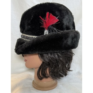 May include: A black faux fur hat with a decorative band and a red feather accent. The hat has a rounded crown and a wide brim. The decorative band is black and white.