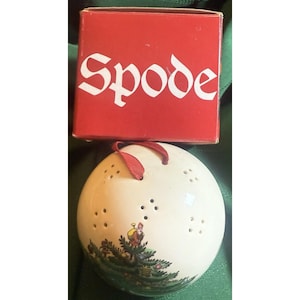 May include: A cream-colored Spode Christmas ornament with a painted scene of a person carrying a sack near a Christmas tree. The ornament has small holes and a red ribbon. The red box above it has the word "Spode" in white.