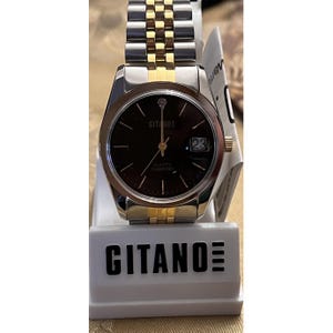 May include: A Gitano quartz diamond watch with a black face, gold and silver band, and date display. The watch is displayed on a white stand with the Gitano logo.