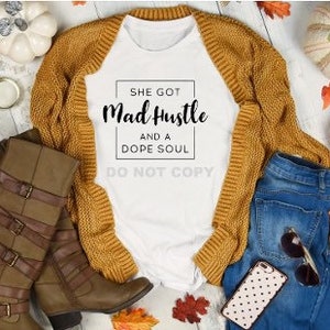 May include: White t-shirt with the text "She got Mad Hustle and a Dope Soul" in a black rectangle. The shirt is styled with a brown cardigan and blue jeans.