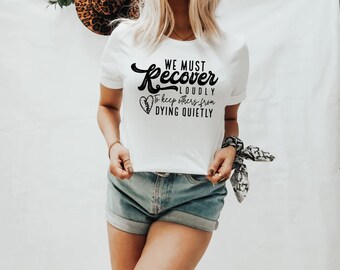 May include: White t-shirt with black text that reads "We must recover loudly to keep others from dying quietly". The text is in a stylized font with a stitched heart graphic. The shirt is being worn by a person with a bandana around their wrist.