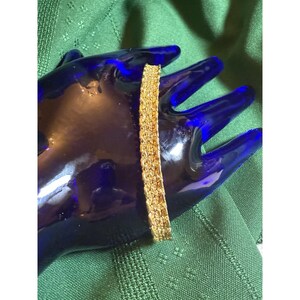 May include: A gold-colored bracelet with a textured surface. The bracelet is displayed on a blue surface against a green background. The bracelet appears to be made of multiple small, connected pieces.
