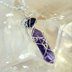May include: A silver wire wrapped pendant featuring a purple amethyst and a black gemstone. The pendant is hanging from a silver chain.
