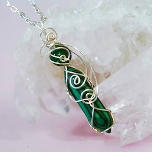 May include: A green malachite stone pendant wrapped in silver wire, hanging from a silver chain. The pendant is displayed on a white crystal background.