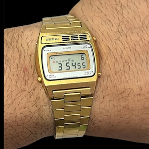 May include: A gold-coloured Seiko digital watch with a rectangular face and a matching link strap. The watch face displays the time, day, and date. The watch is on a wrist.