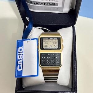 Może przedstawiać: Złoty zegarek Casio Data Bank z funkcją kalkulatora. Zegarek znajduje się w oryginalnym pudełku z etykietą Casio.