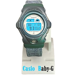 May include: A Baby-G digital watch with a blue digital display showing the date and time. The watch has a green and gray case and a patterned gray strap. The watch is displayed on a white stand with the text "Casio Baby-G".