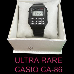 Op de afbeelding: Een zwarte Casio CA-86 rekenmachine-horloge in een open witte doos met een zacht, wit kussen. Het horloge heeft een digitaal display en knoppen. De tekst "ULTRA RARE CASIO CA-86" is eronder te zien. Een vintage digitaal horloge.