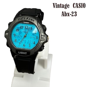 May include: Vintage Casio Abx-23 digital watch with a black strap and a light blue face. The watch features a world map design and the text "DATA BANK" and "LIGHT". The watch is displayed on a white stand.