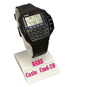 Op de afbeelding: Een zwarte Casio Cmd-20 rekenmachine horloge op een witte display stand. Het horloge heeft een digitaal display met tijd en datum, en een toetsenbord. De display stand heeft de woorden "RARE" en "Casio Cmd-20" erop gedrukt.