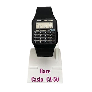 May include: A black Casio CA-50 digital watch with a calculator-style keypad. The watch displays the time as 5:10:25 AM. The watch is on a white display stand with the word "Rare" and "Casio CA-50" printed on it.