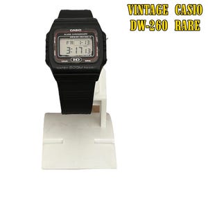 May include: A black Casio DW-260 digital watch with a rectangular face, displaying the date and time. The watch is on a white display stand. The text "VINTAGE CASIO DW-260 RARE" is in the upper right corner.