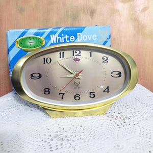 May include: A gold-colored oval alarm clock with a white face and black numbers. The clock has a white dove logo and the words "White Dove" printed on the face. The clock is sitting on a white crocheted surface.