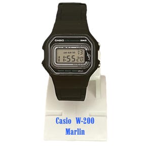 Puede incluir: Reloj digital Casio W-200 Marlin negro. El reloj muestra la hora 11:55:20 y la fecha 13. Tiene una correa negra y una esfera blanca y negra. El texto "WATER RESIST 100M" es visible. El reloj está sobre un soporte blanco.