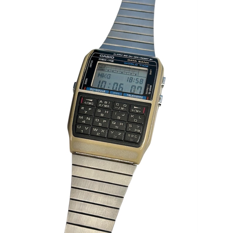 May include: A vintage Casio DBX-118 digital watch with a gold-tone case and a silver-tone metal band. The watch face displays the time and features a calculator keypad. The watch shows the time as 10:06:07 and the city code HKG.