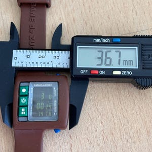 May include: A brown Cabane de Zucca digital watch with a rectangular face, displaying the time and date. The watch is being measured with a digital caliper, showing a width of 36.7 mm. The watch band is also brown.