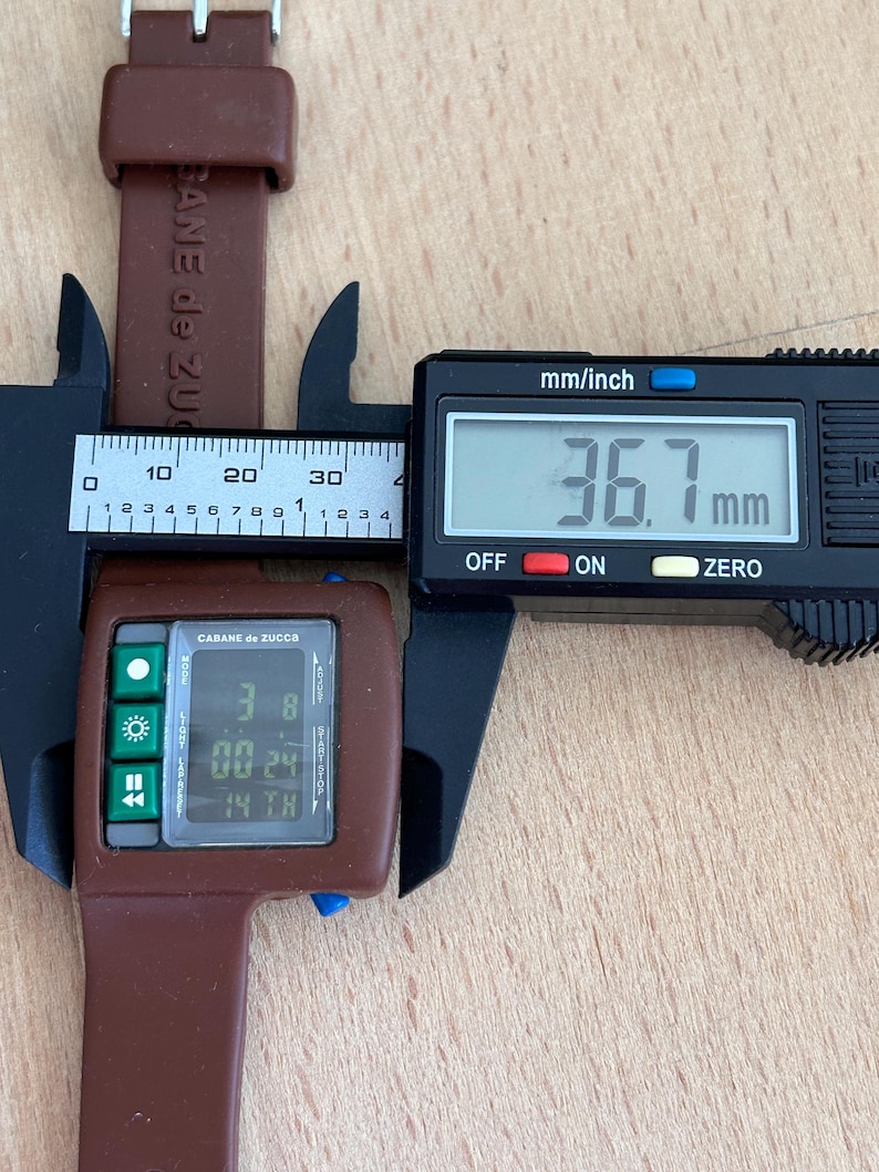 May include: A brown Cabane de Zucca digital watch with a rectangular face, displaying the time and date. The watch is being measured with a digital caliper, showing a width of 36.7 mm. The watch band is also brown.