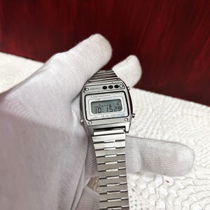 Puede incluir: Un reloj digital Seiko plateado con una correa de metal plateado. La esfera del reloj muestra la hora, la fecha y el d&iacute;a de la semana. El reloj tiene una pantalla digital negra con n&uacute;meros blancos. El reloj tiene un bot&oacute;n negro en la parte superior derecha de la esfera.